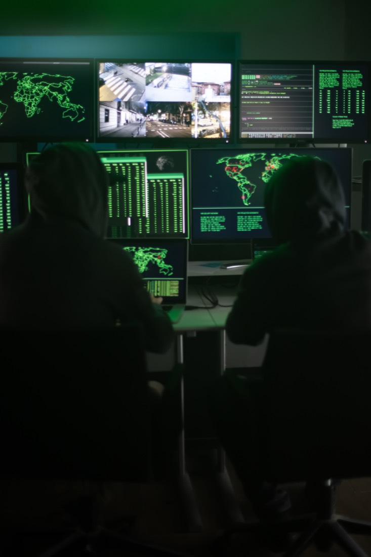 Two hooded figures working at screens in a dim basement.